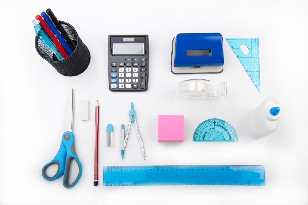 back-to-school essentials including pens, scissors, a calculator, a ruler, protractor, compass, and glue on a white table