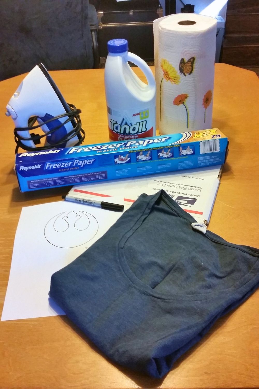 A navy t-shirt, paper rebel stencil, freezer paper, marker, iron, cardboard box, bottle of bleach, and roll of paper towels.