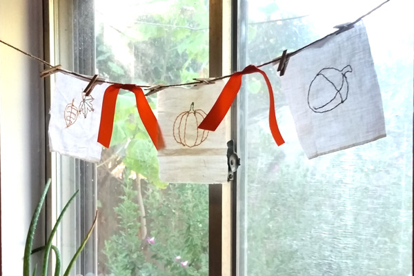 embroidered bunting with a square of leaves, a pumpkin, and an acorn strung across a window on jute