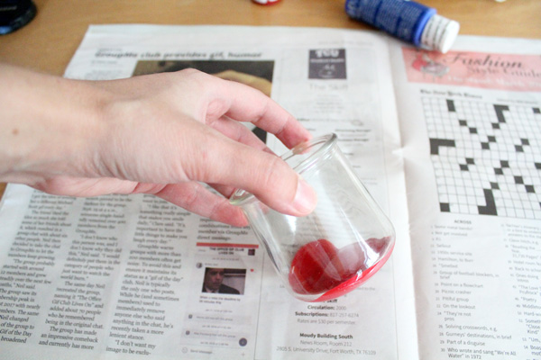 A hand holding a glass jar to swirl red paint inside.