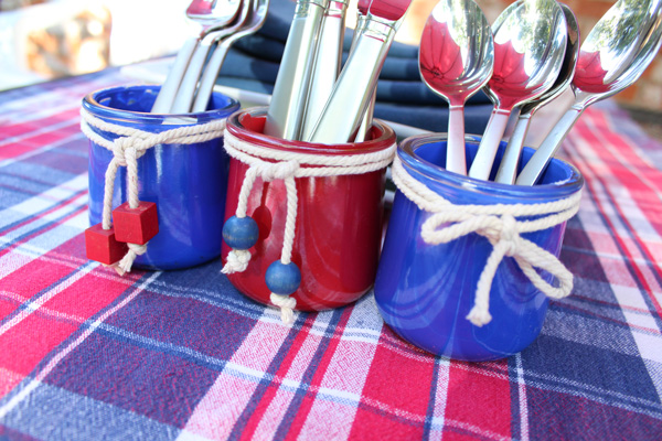 Three painted jars holding silver forks, knives, and spoons on a plaid tablecloth.