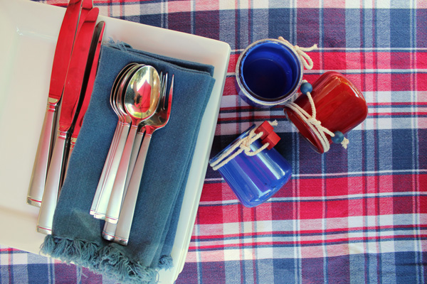 Overhead view of silverware, blue napkins, white plates, and painted silverware holders on a red, white, and blue plaid tablecloth.