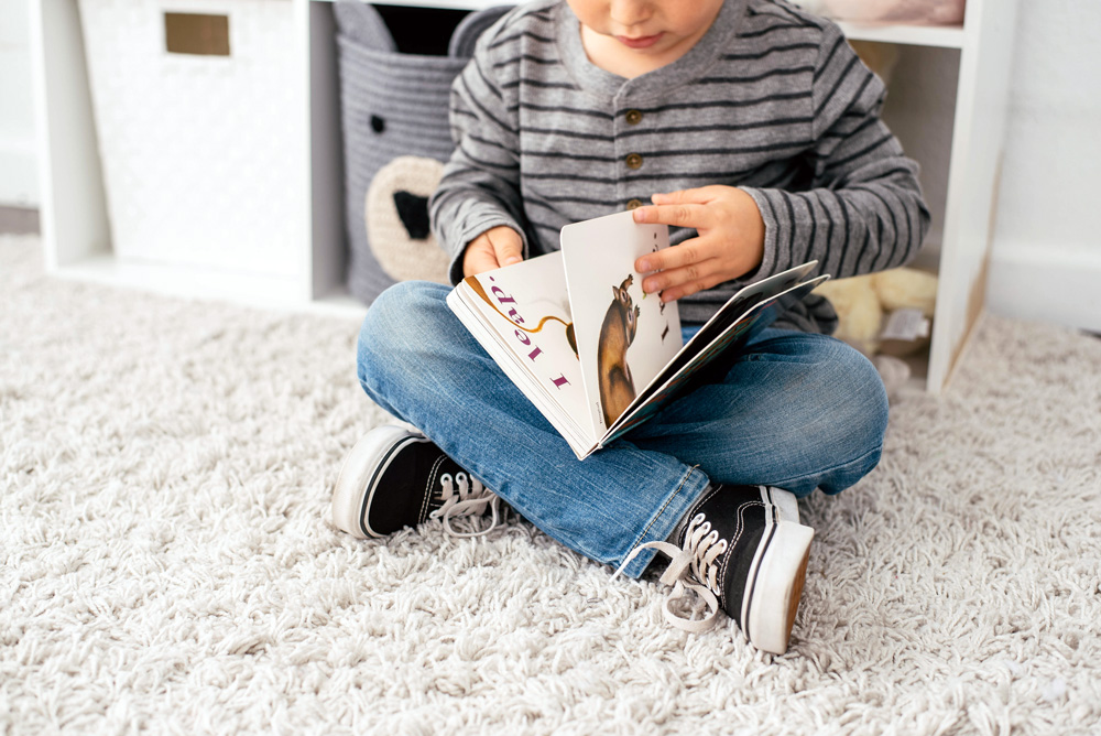 Young child reading a board book on a carpet floor.