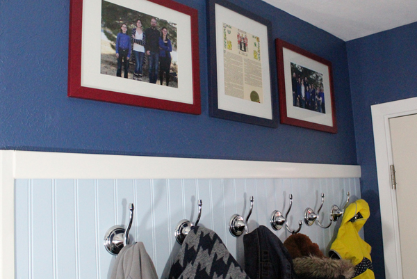 Coats hang from chrome hooks on a blue paneled wall. Above the coats, a darker blue wall has 3 frames photos.