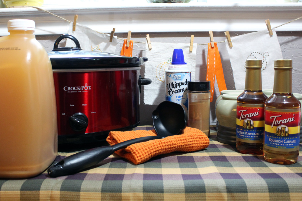 table with a slow cooker and a jug of cider, can of whipped cream, bottles of Torani syrup, and a big ladle