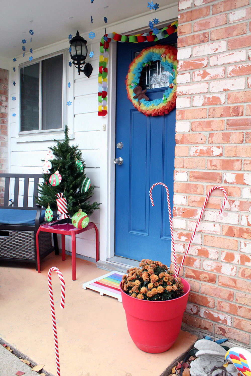 front porch with orange brick, white siding, and a blue door decorated with a rainbow wreath and garland