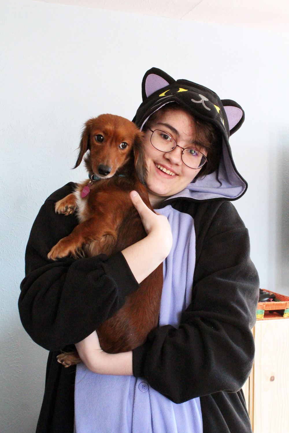 child wearing a black cat kigurumi and holding a red dachshund