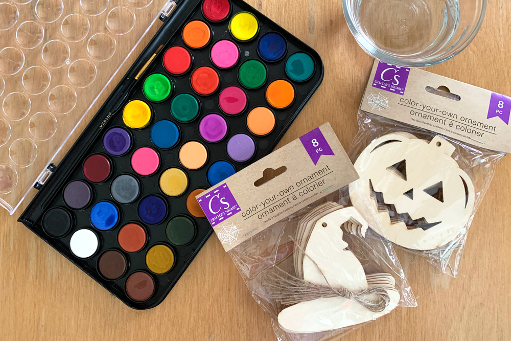 Watercolor paint kit with a brush, a cup of water, and packages of wooden hat and jack o'lantern ornaments.