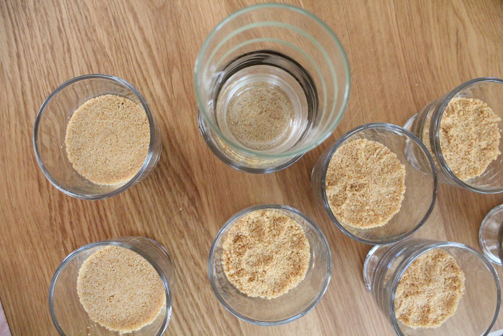 Pressing graham cracker crust mixture into the bottom of several glasses with another glass.