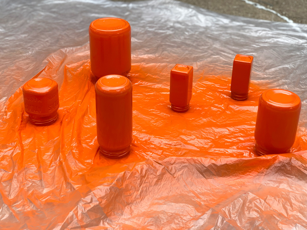Several glass jars upside down on a plastic sheet and spray painted orange.
