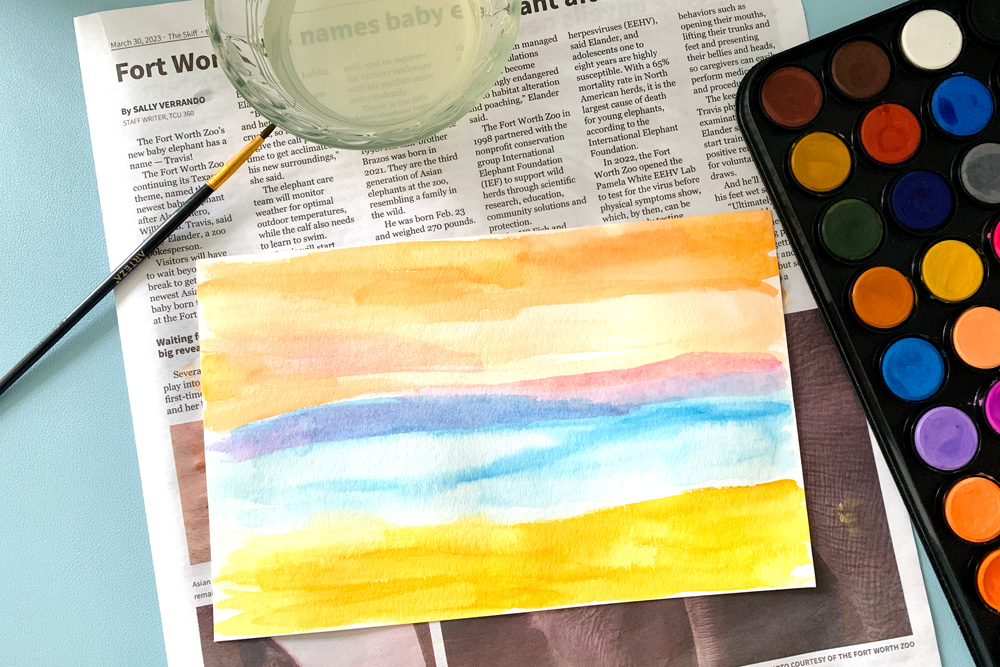 Painting a watercolor landscape background in orange, pink, blue, and golden brown.
