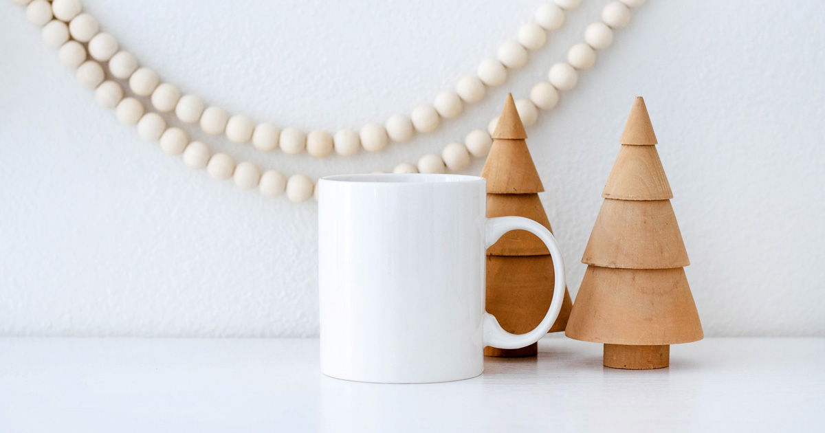 Blank white mug beside 2 small wood Christmas tree and a bead garland.