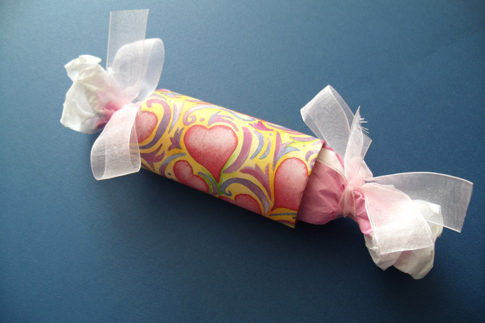 A paper tube wrapped in pink heart paper and filled with tissue paper tied on the ends to resemble a cracker.