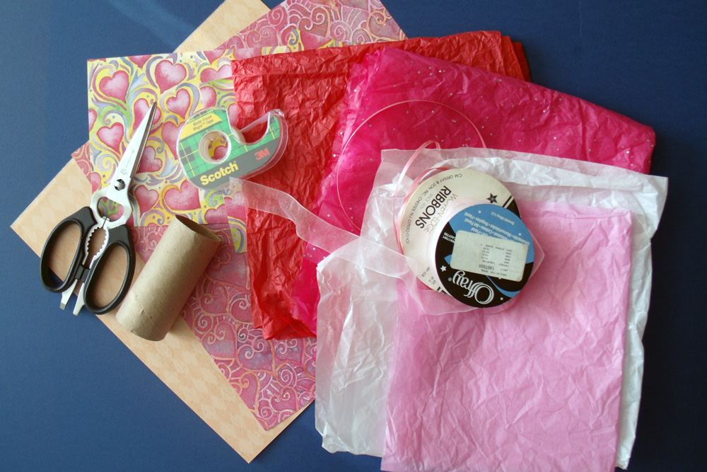 Heart-patterned scrapbook paper, pink and red tissue paper, a paper tube, pink ribbon, scissors, and a roll of tape.