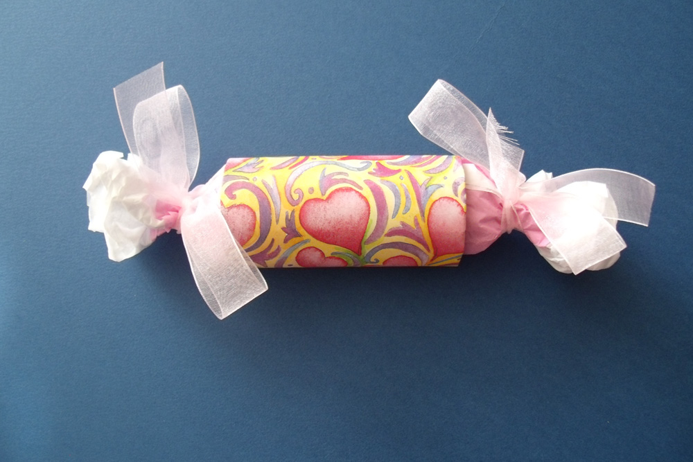 A paper tube wrapped in pink heart paper and filled with tissue paper tied on the ends to resemble a cracker.