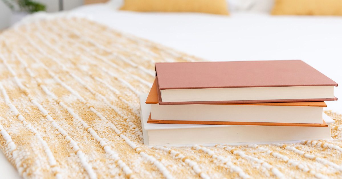 A stack of 3 orange hardback books on a yellow bed spread.