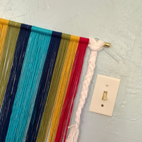 DIY rainbow wall hanging made from red, yellow, green, navy, and light blue yarn and a white braid.