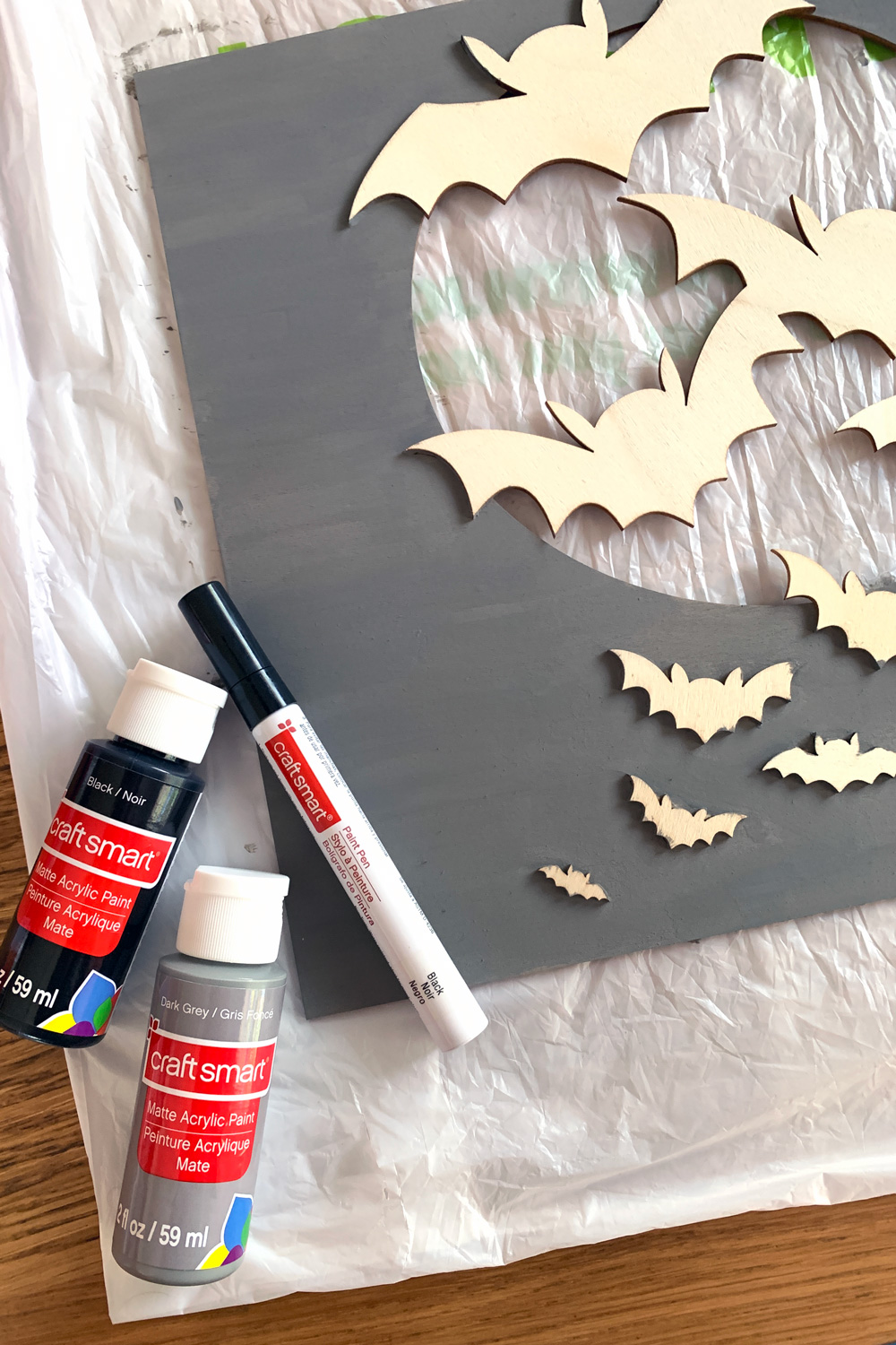 Painting the background of the wood sign gray beside containers of black and gray paint and a black paint pen.