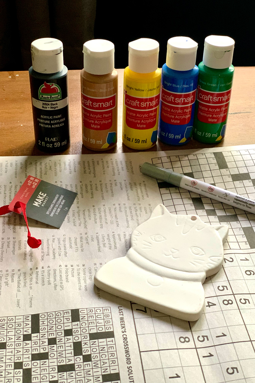 An unpainted white cat ornament on a newspaper beside several bottles of craft paint.