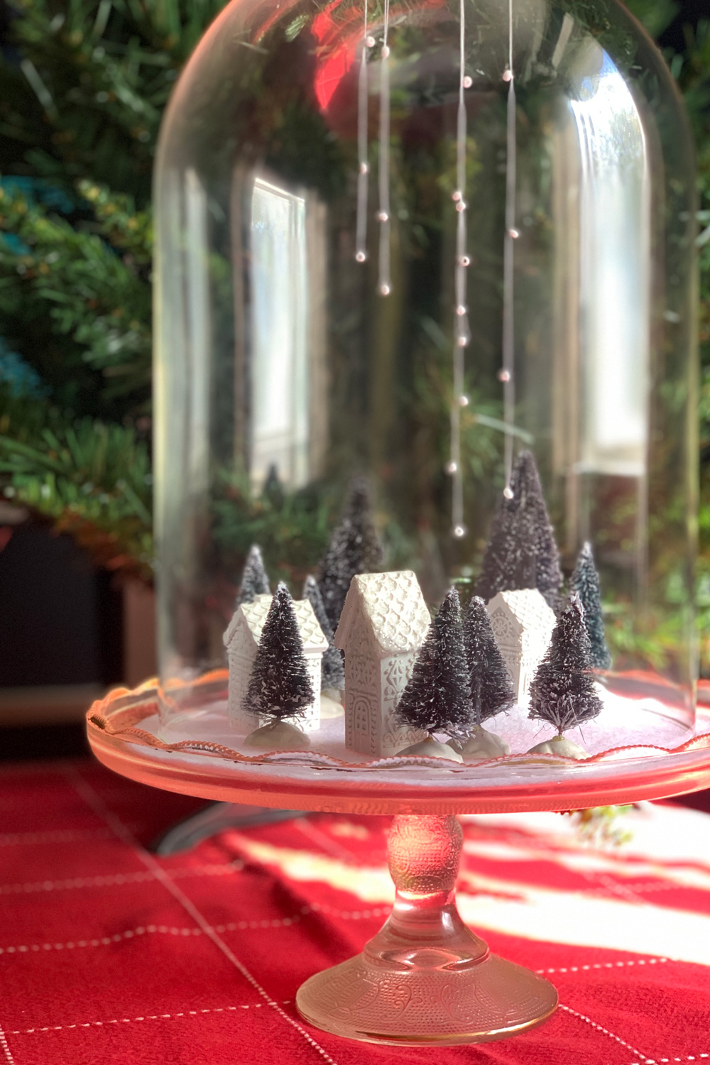 Christmas village with tiny white houses and green trees on white felt snow under a glass cloche dome.