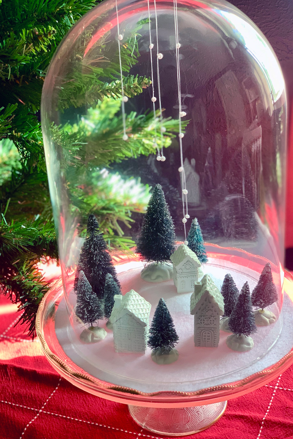 A village of white houses and green trees under a glass cloche dome with bead snowflakes hanging from the top.