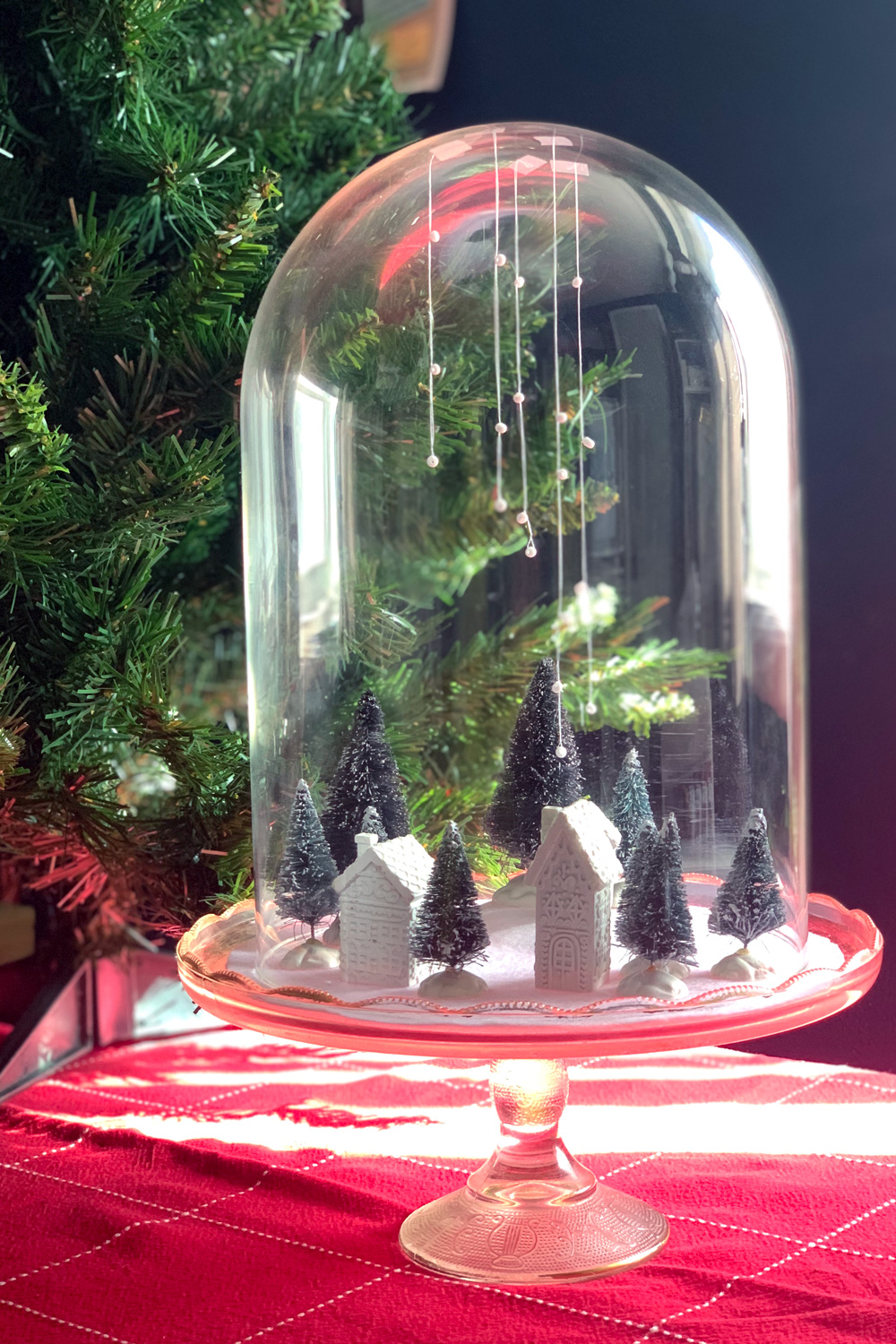 Christmas village with tiny white houses and green trees on white felt snow under a glass cloche dome.
