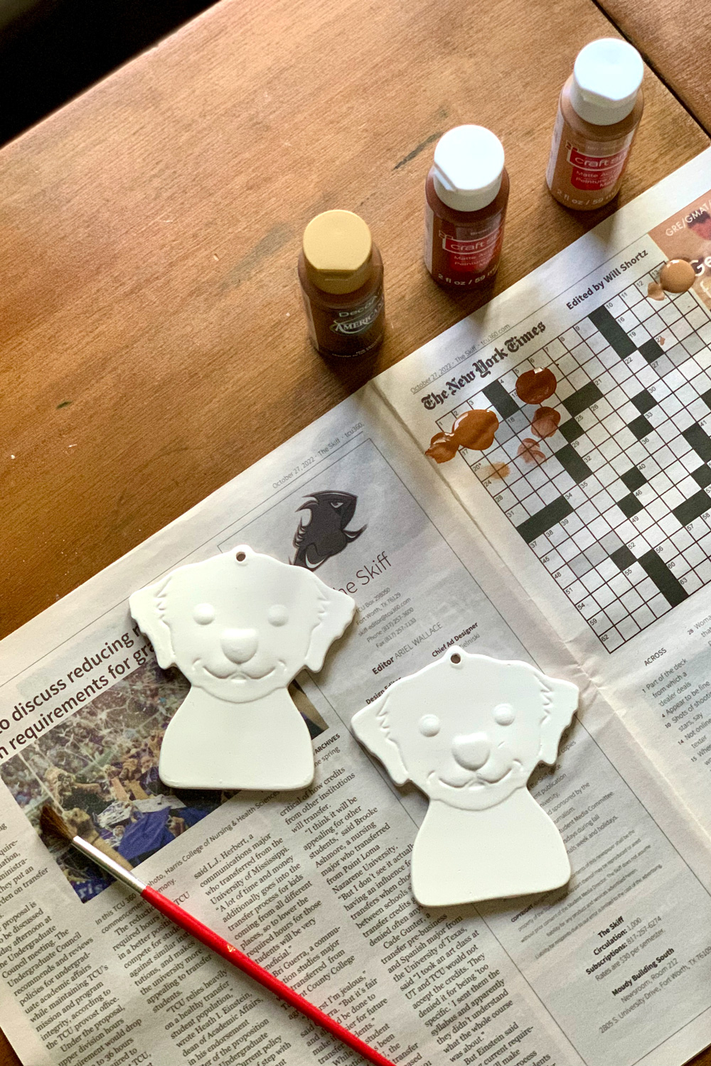 Two unpainted white dog ornaments on a newspaper beside several bottles of craft paint.