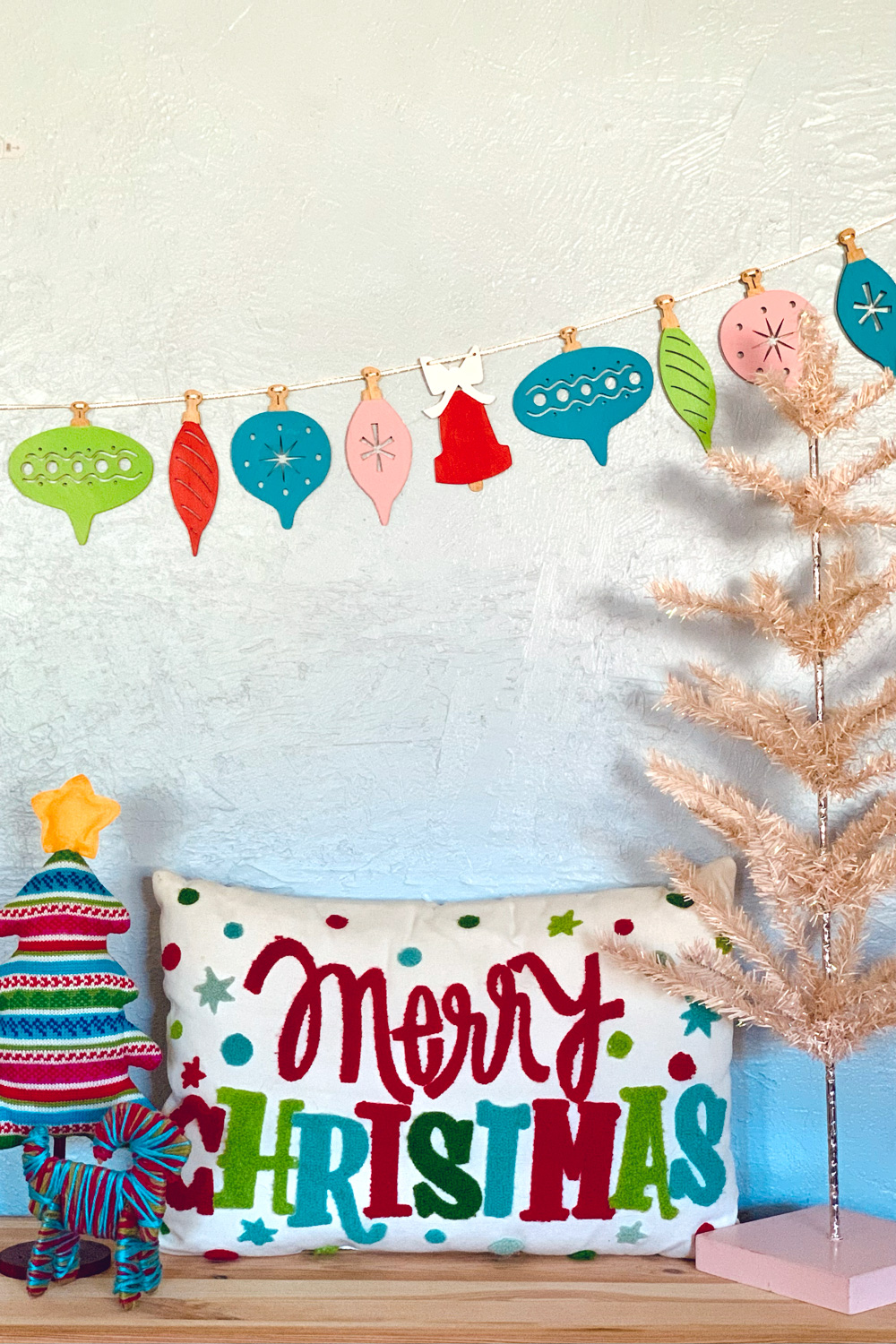 Retro wood ornament garland in pink, red, teal, and lime green beside matching Christmas pillow and trees.