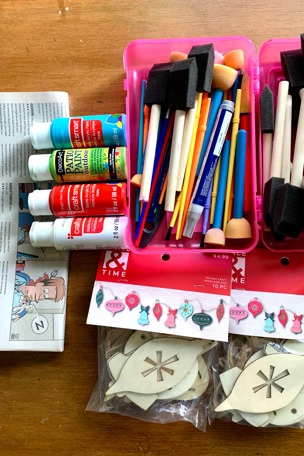 Newspaper, paint bottles, a box of paintbrushes, and 2 packaged of unfinished wood ornaments.