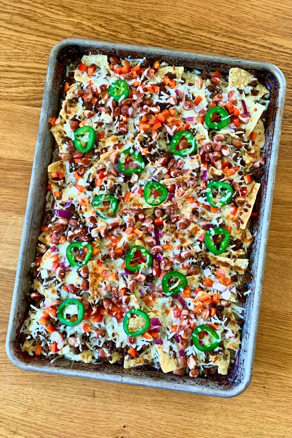 A sheet pan filled with chips, meat, beans, onion, jalapeno slices, and melted cheese.