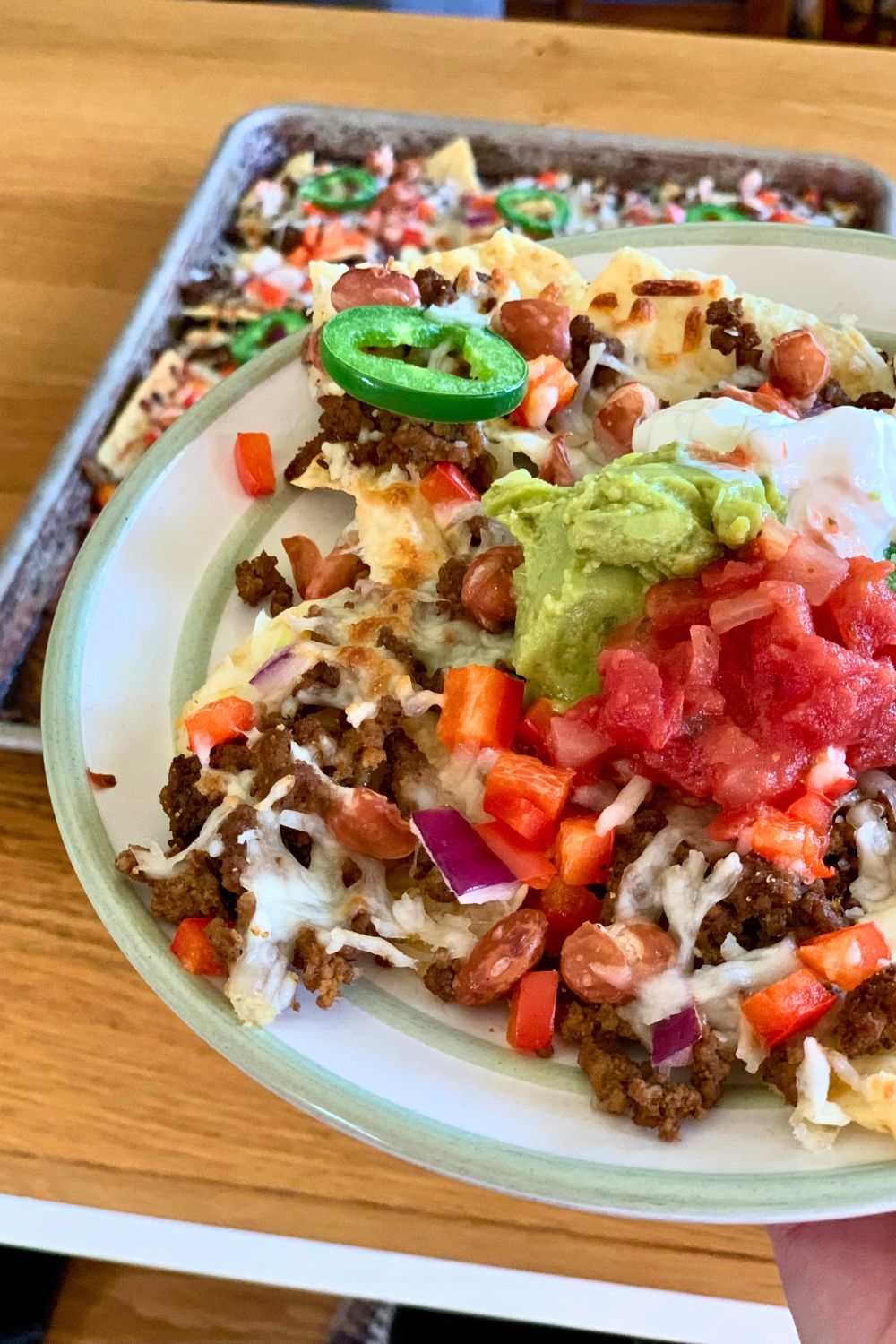 A hand holding a loaded plate of nachos above a sheet pan fresh from the oven.