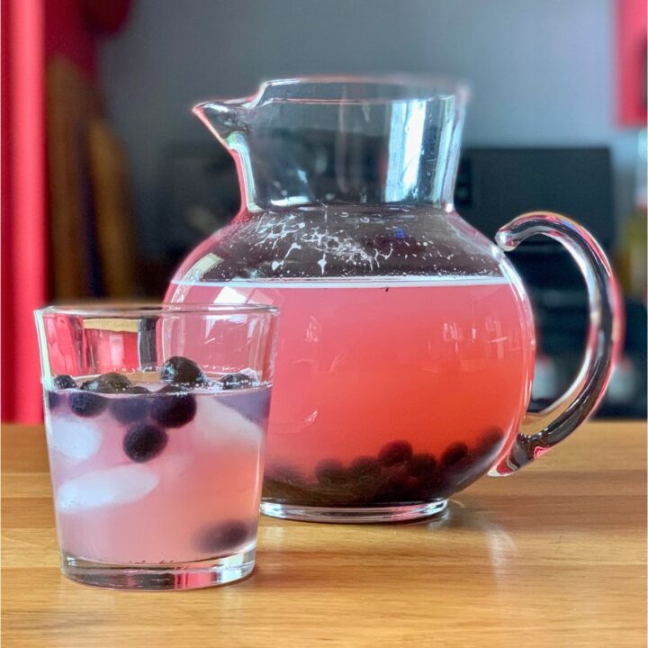 A glass and pitcher of pink honey blueberry lemonade with fruit.