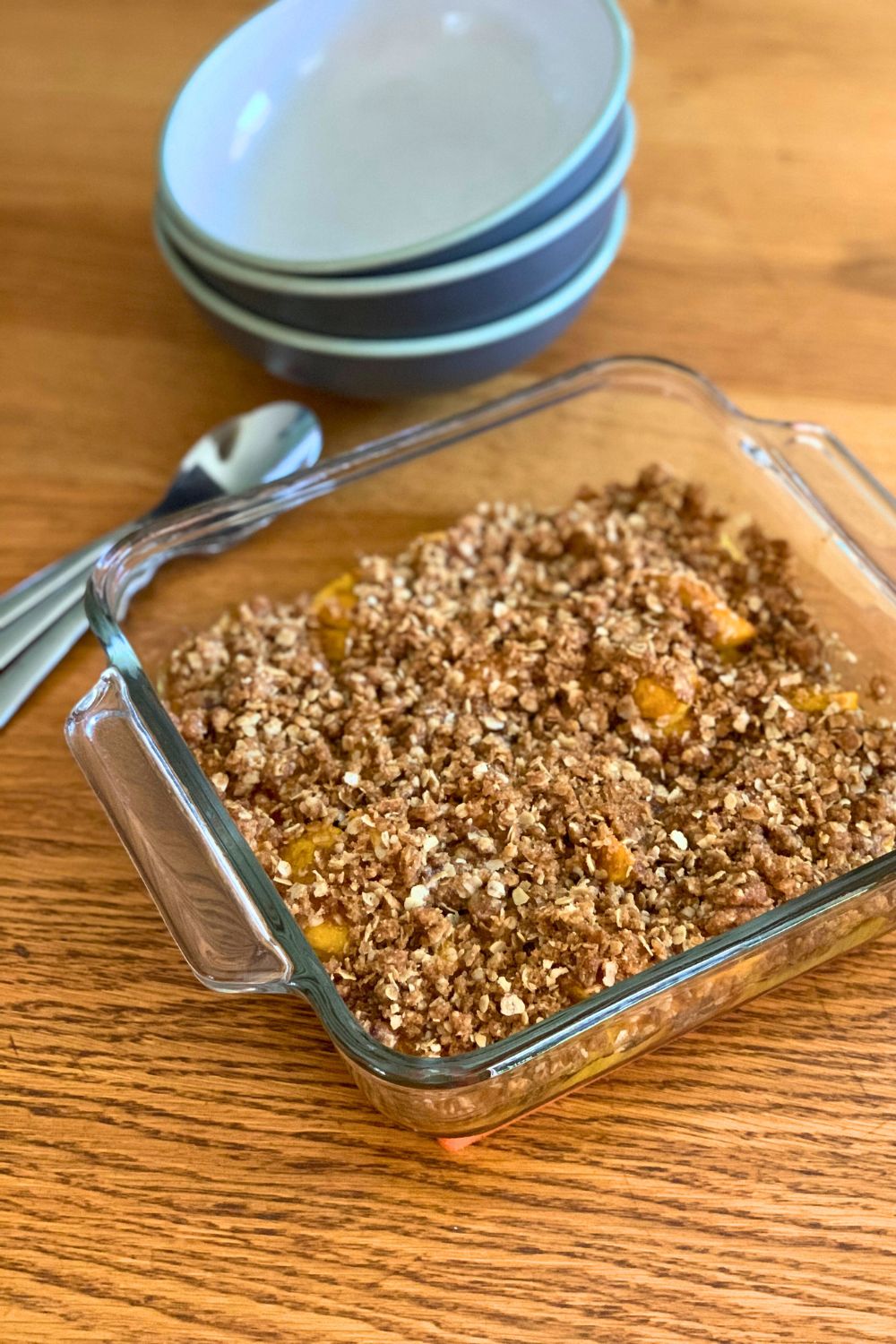 Peach fruit crisp in a baking dish beside a stack of bowls and spoons.