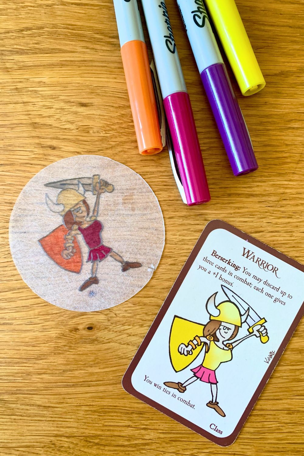 A Munchkin game card with a warrior girl beside a circle of Shrinky Dink plastic with a traced image colored pink and orange.