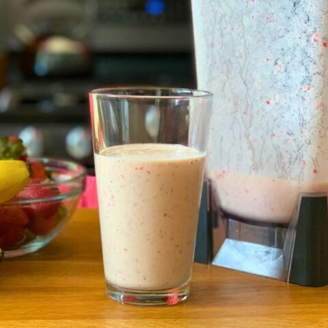 Orange sunrise smoothie in a glass beside a blender, a bunch of bananas, and a bowl of strawberries.