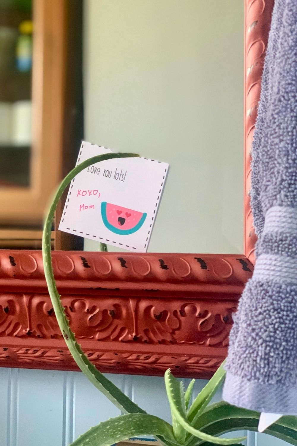 A bathroom mirror with a white notecard stuck to the edge that says 'love you lots' signed XOXO Mom beside a heart-eyed watermelon.