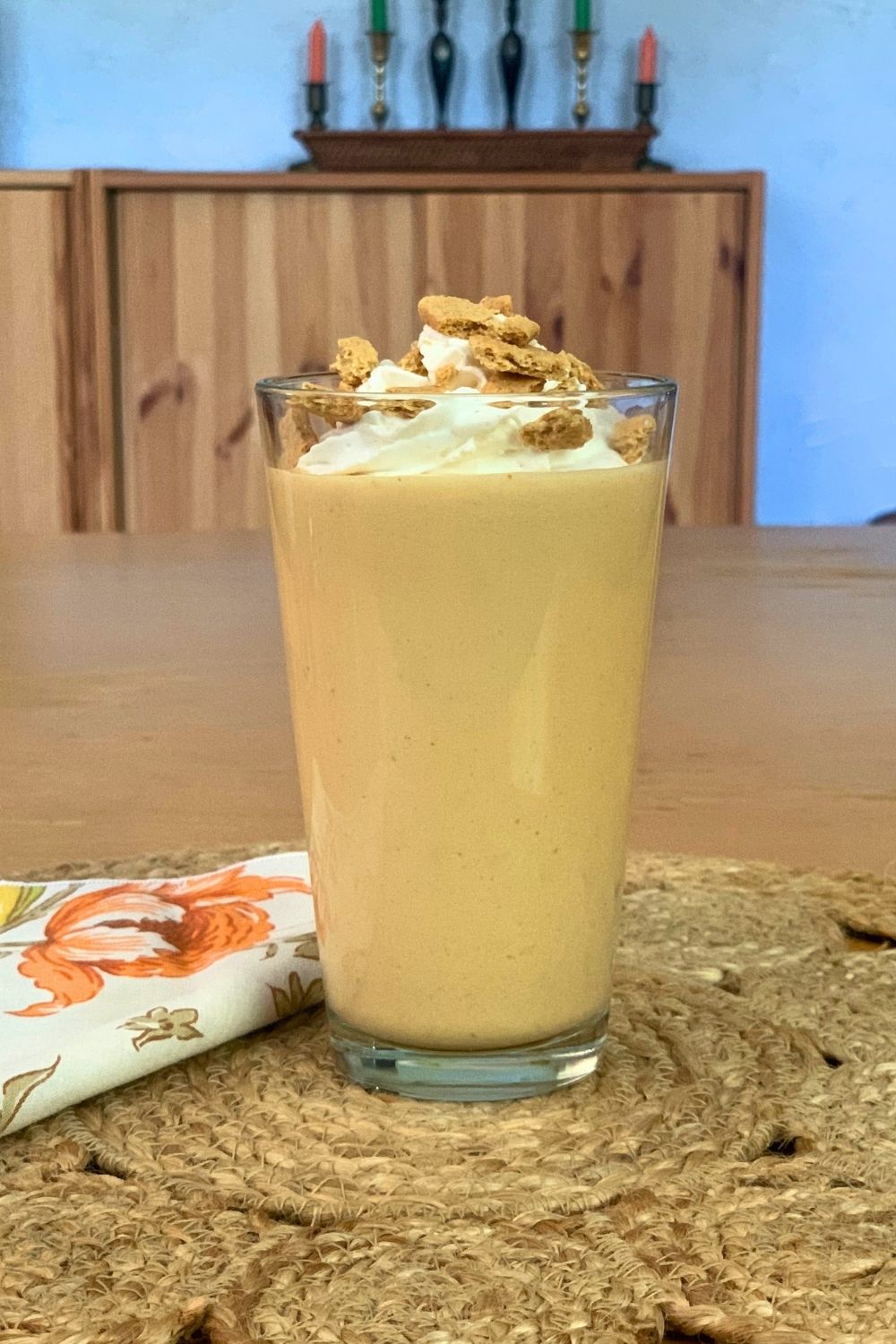 A glass filled with a golden orange pumpkin smoothie topped with whipped cream and graham crackers.
