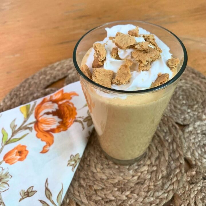 A glass filled with a golden orange pumpkin smoothie topped with whipped cream and graham crackers.