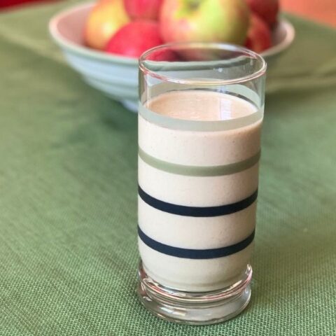An apple pie smoothie in a glass beside a bowl of fruit.