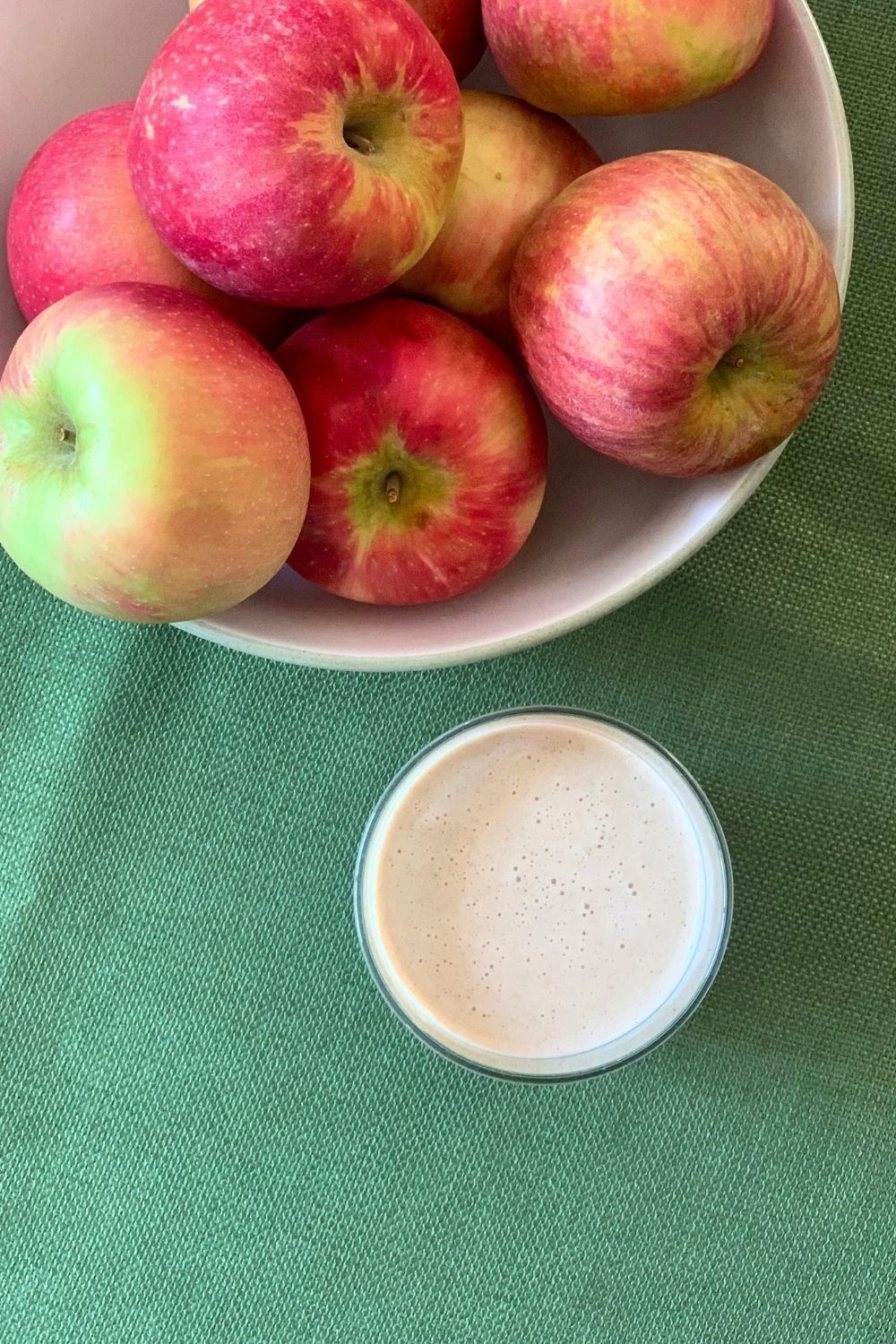 Overhead view of a thick off-white drink beside a bowl of pink-red apples