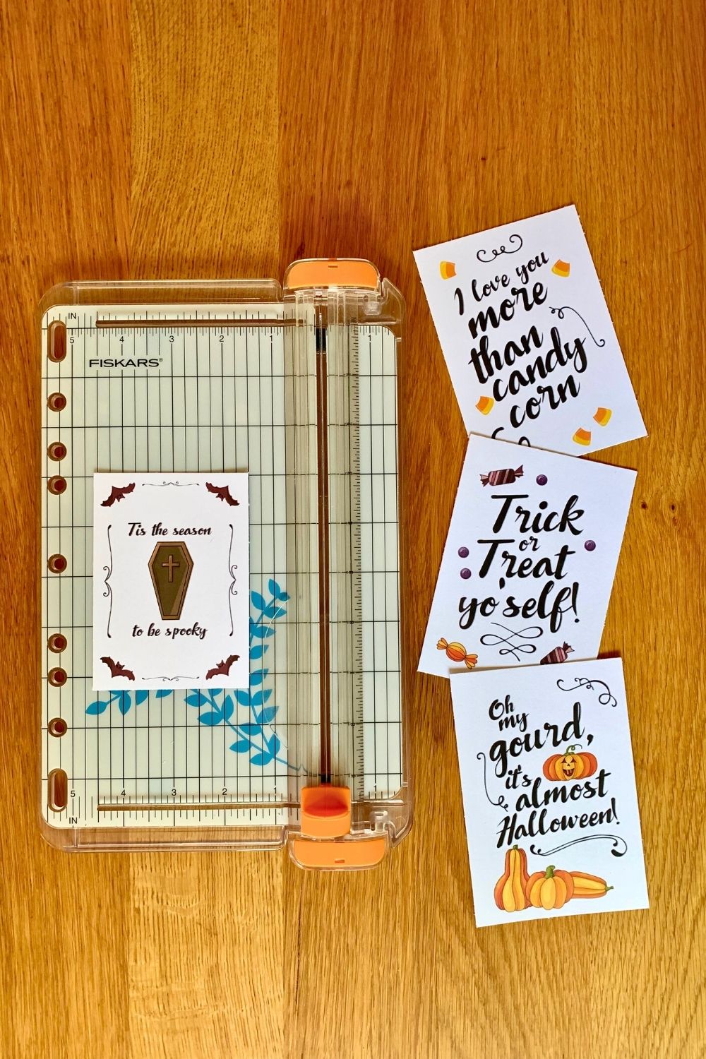 A paper trimmer beside cards that read "tis the season to be spooky," "I love you more than candy corn," "trick or treat yo'self," and "Oh my gourd it's almost Halloween."