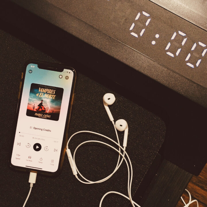 Vampires of El Norte audiobook via Audible on a phone screen beside earbuds on a walking pad.