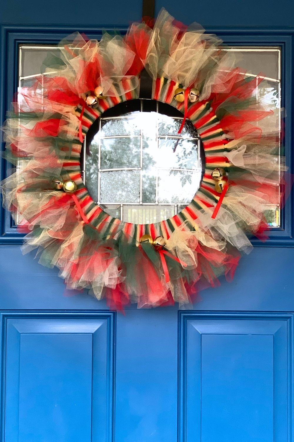 Red, gold, green, and silver tulle Christmas wreath with jingle bells on a bright blue door.