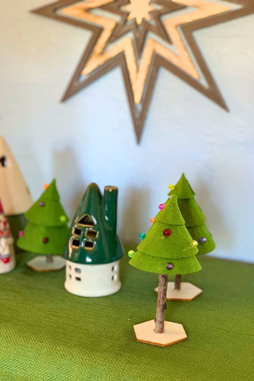 A Christmas tree craft on a green tablecloth beside a ceramic house.