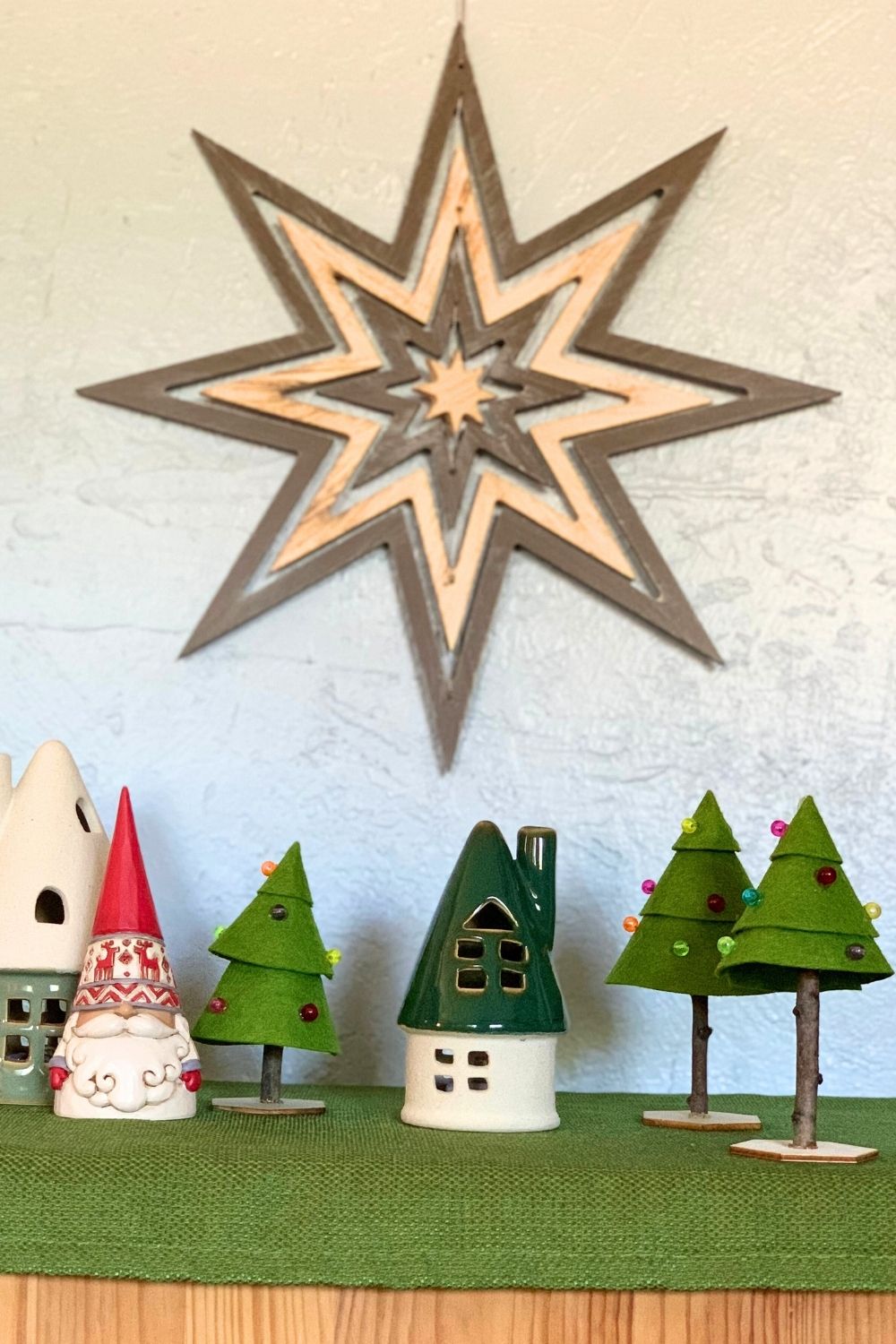 Handmade felt Christmas trees on a table display beside small ceramic houses and a gnome figure.