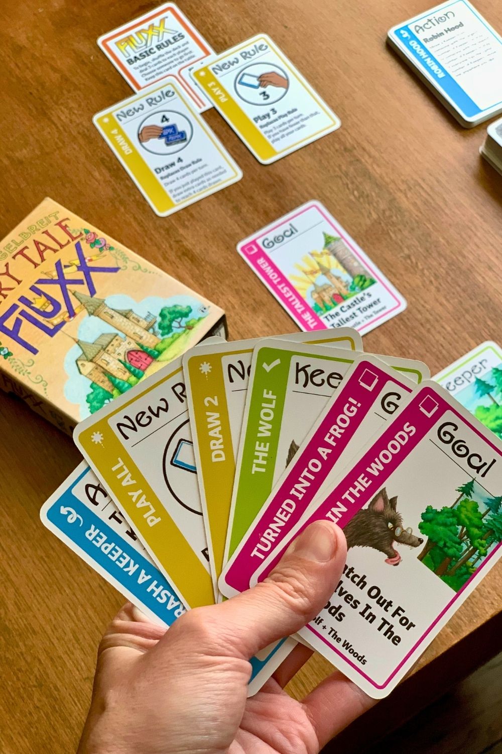 A hand holding half a dozen Fairytale Fluxx cards with the game set up on the table in the background.