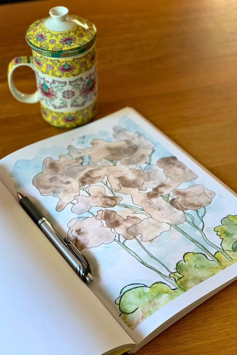 A pen sitting in the seam of an open Reverse Coloring Book with a blue, green, and brown page outlined to resemble a stand of trees.