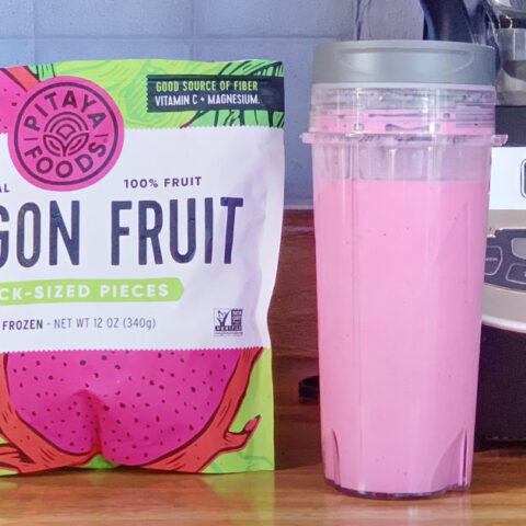 A bright pink dragonfruit smoothie in a plastic tumbler beside a bag of frozen fruit.
