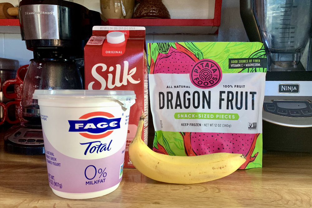 A carton of yogurt, container of soy milk, banana, and bag of dragon fruit on a kitchen counter.