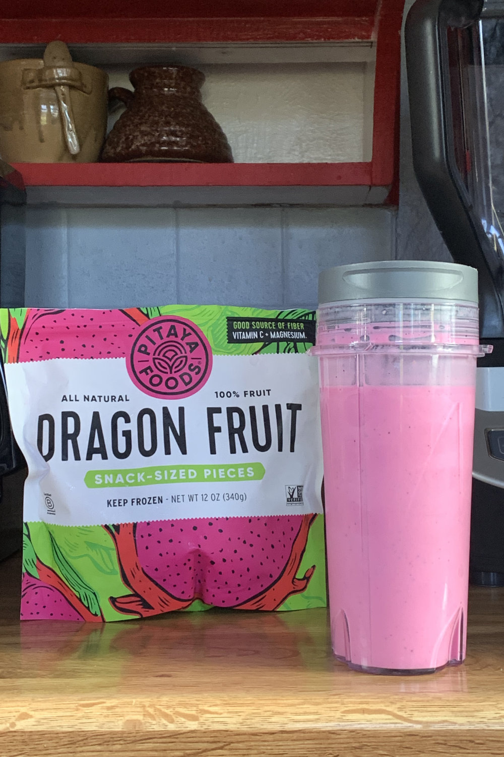 A bright pink dragonfruit smoothie in a plastic tumbler beside a bag of frozen fruit.